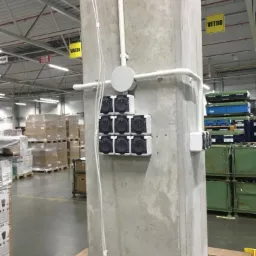 Betonowy słup w hali magazynowej z zamontowanymi gniazdami elektrycznymi w białych obudowach, okablowaniem w białych rurkach i przewodami, widoczne palety i regały w tle.