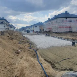 Budowa osiedla domów jednorodzinnych w trakcie realizacji, widoczne fundamenty, nasypy ziemi, ubijarka, oraz budynki w trakcie ocieplania z reklamą materiałów izolacyjnych.