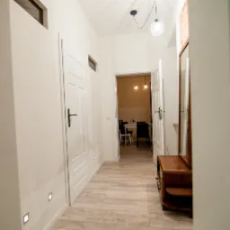 Jasny korytarz z białymi ścianami, drewnianą podłogą i oświetleniem punktowym. Widoczne drzwi, lustro w drewnianej ramie i wnętrze pokoju z jadalnią w tle.