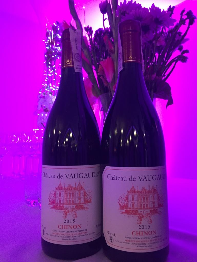 Dwie butelki czerwonego wina Château de Vaudgaudry Chinon 2015 na stole, z fioletowym oświetleniem i kwiatami w tle.