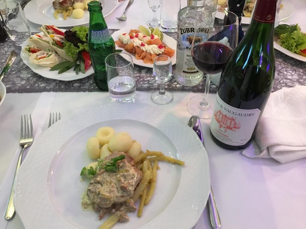 Uroczysty stół z różnorodnymi daniami, butelką wina Château de Vaudgaudry Chinon 2015, wódką Żubrówka i napojem w zielonej butelce, nakryty białym obrusem z szarym bieżnikiem.