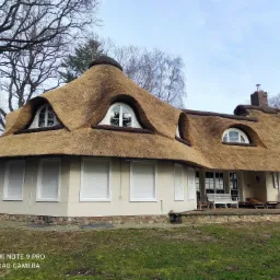 Dom z charakterystycznym strzechą, widoczne okna dachowe i fragment elewacji z jasnym tynkiem i kamienną podmurówką, drzewa bez liści w tle.