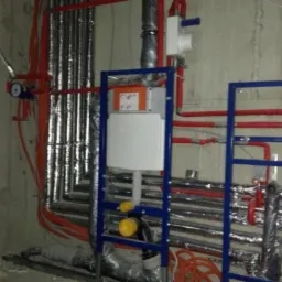 Instalacja podtynkowa WC w trakcie montażu: widoczne rury w izolacji, stelaż podtynkowy w kolorze niebieskim, oraz część instalacji wodnej w kolorze czerwonym i niebieskim.