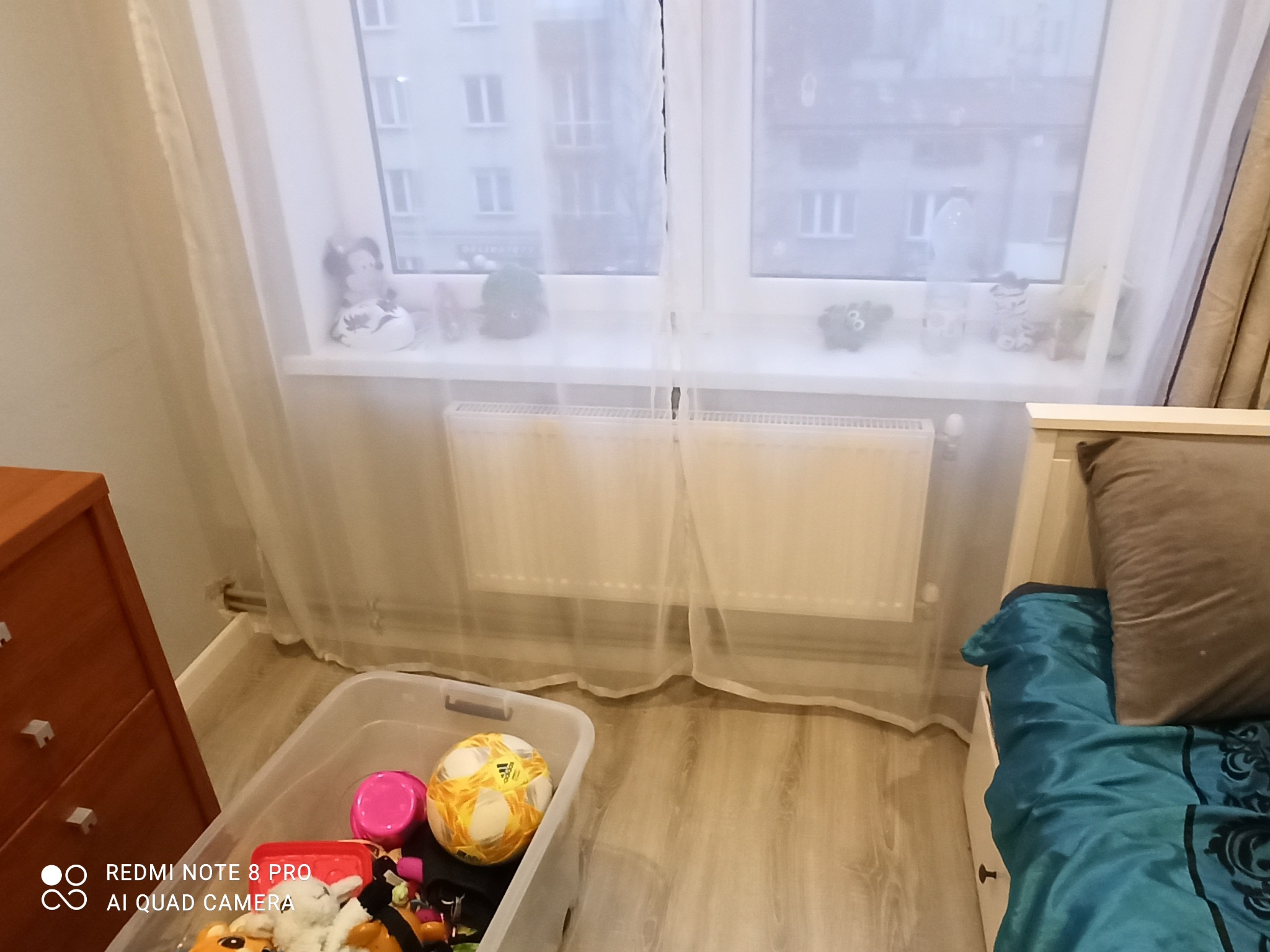 Pokój dziecięcy z grzejnikiem pod oknem, obok komoda i łóżko, w rogu przezroczysty pojemnik z zabawkami.