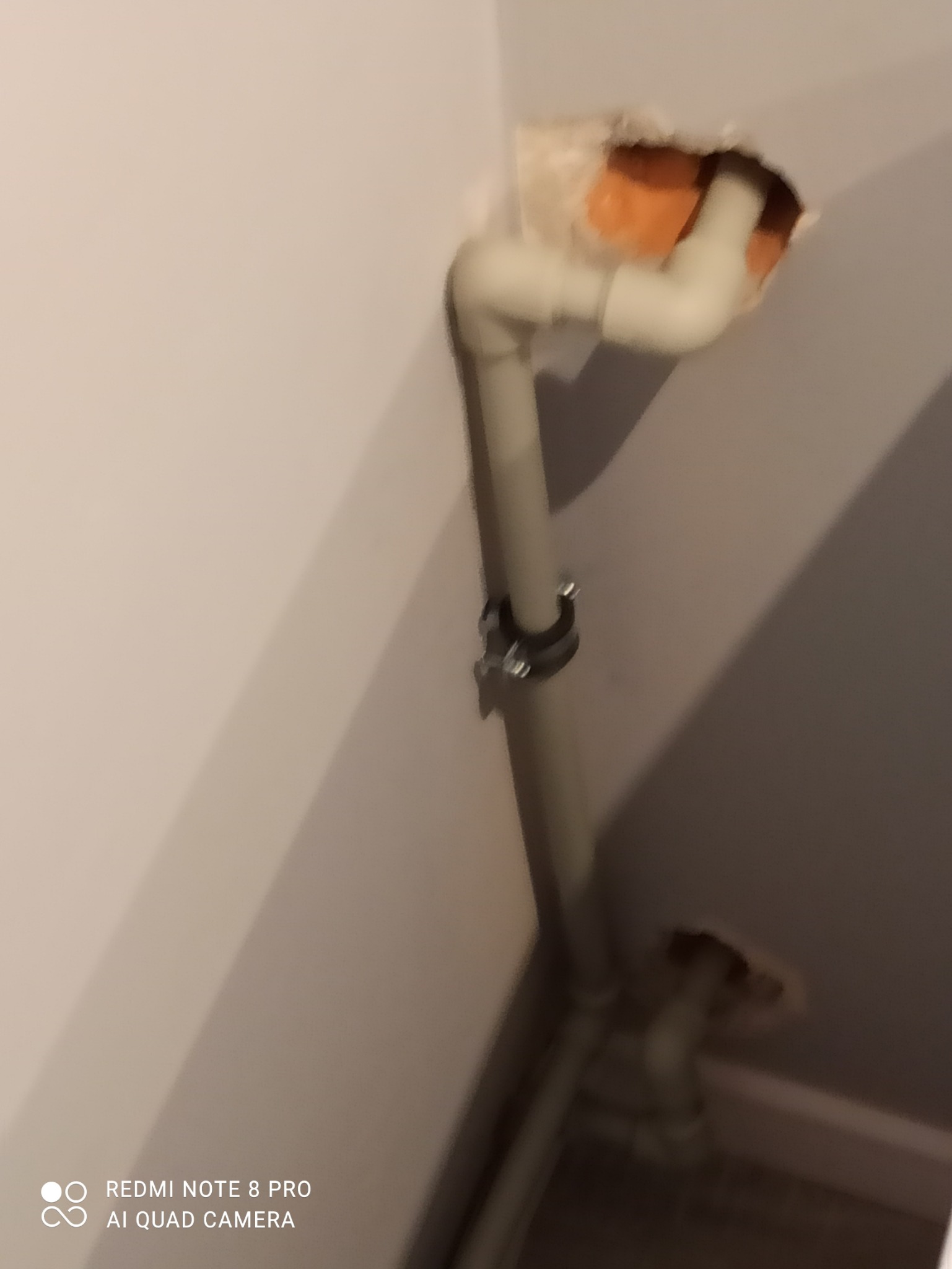 Niedokończona instalacja rur w łazience, widoczna dziura w ścianie z pomarańczową izolacją, rury przymocowane obejmą do ściany.