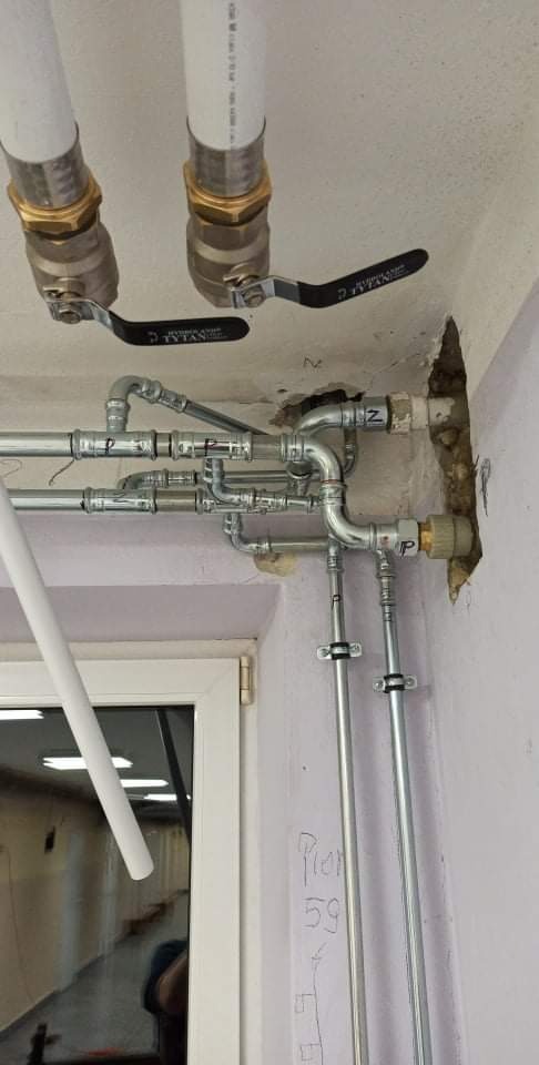 Widok na sufit z nową instalacją wodną z rur PCV i metalowych, z zaworami Tytan, w trakcie remontu łazienki, widoczne ślady po kuciu w ścianie, instalacja częściowo nad oknem.