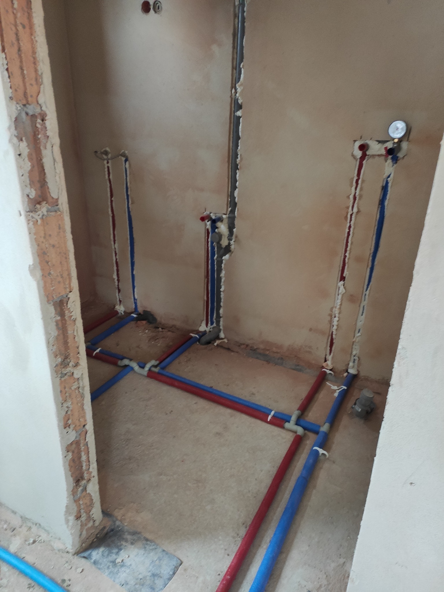 Wnętrze łazienki w trakcie prac instalacyjnych: widoczne rury wodne (czerwone i niebieskie) prowadzone w ścianach i podłodze, zabezpieczone pianką montażową, ściany w stanie surowym.