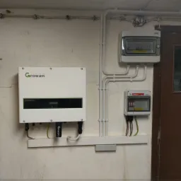 Instalacja fotowoltaiczna w piwnicy: inwerter Growatt, rozdzielnica elektryczna i zabezpieczenia Edison zamontowane na ścianie z widocznymi przewodami.