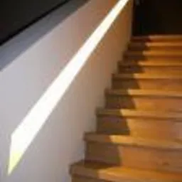 Klatka schodowa z drewnianymi schodami oświetlona promieniem słońca padającym na gładką, białą ścianę. Widoczne ciemne tło w górnej części kadru.