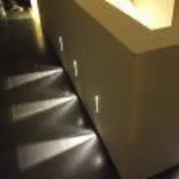 Ciemna podłoga z połyskliwymi płytkami oświetlona punktowymi lampami wbudowanymi w białą ścianę, tworzącymi geometryczne wzory światła.