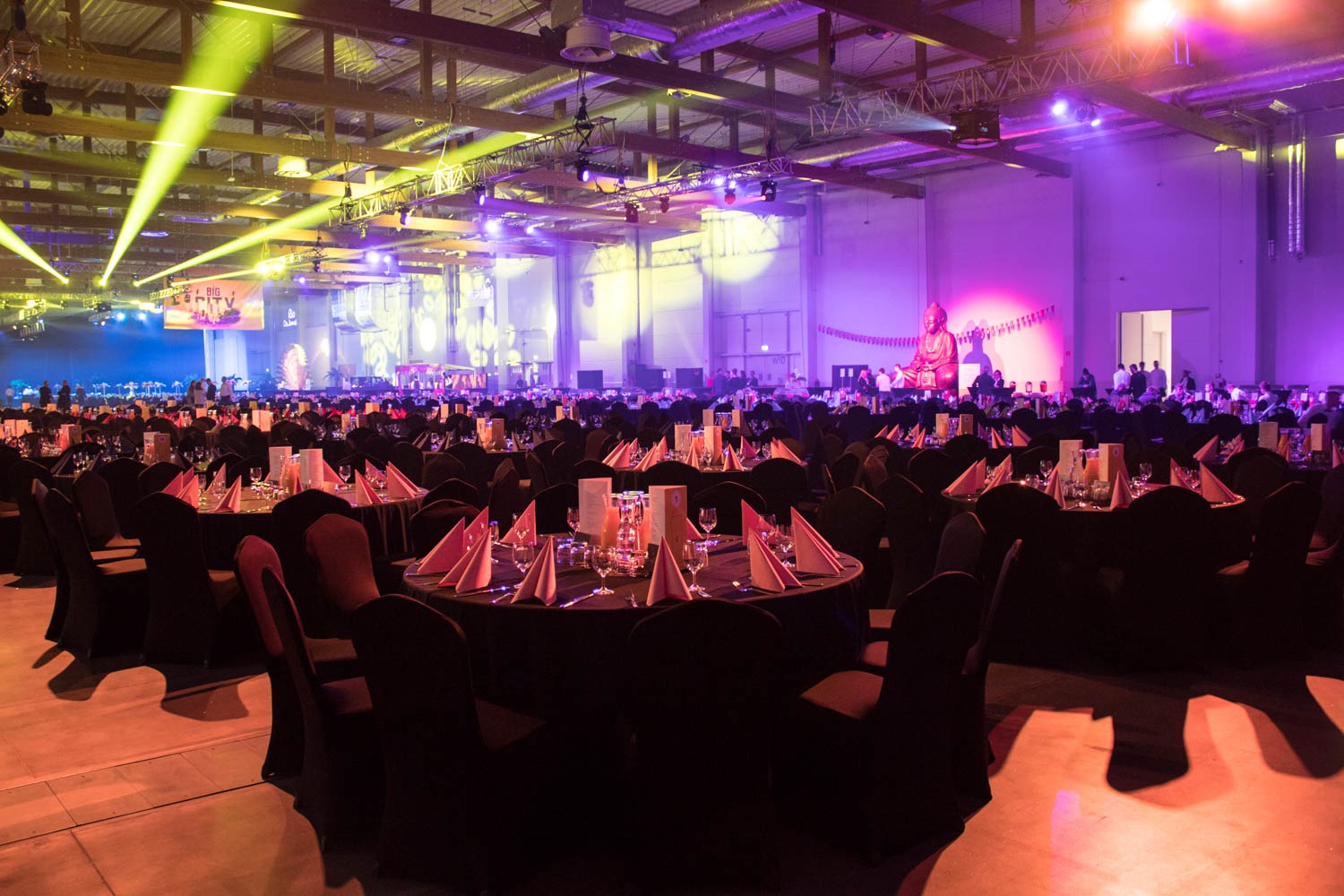 Sala bankietowa z okrągłymi stołami nakrytymi czarnymi obrusami, różowe serwetki, szkło, oświetlenie sceniczne, w tle scena z dekoracją i posągiem Buddy.