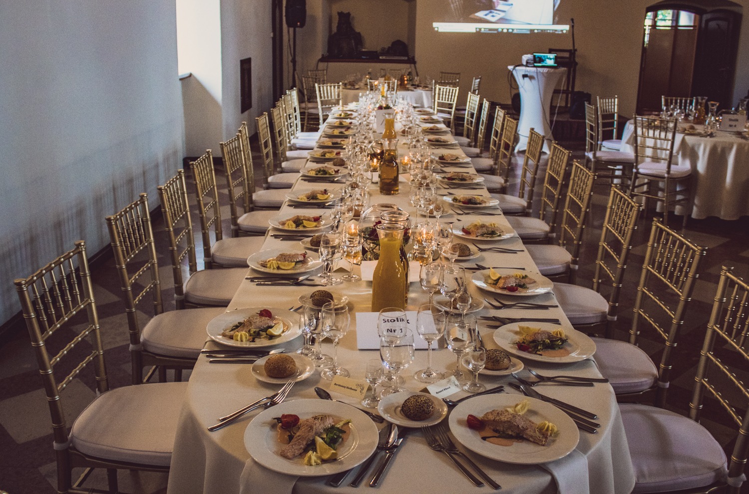Długi stół bankietowy z białym obrusem, nakryty talerzami z rybą, pieczywem, szklankami i karafkami z napojem, ustawiony w sali z krzesłami w kolorze złota.