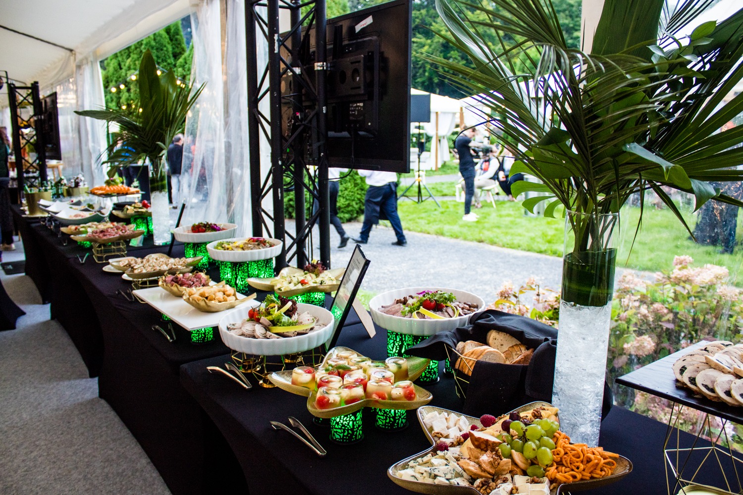 Stół cateringowy z różnorodnymi przekąskami i daniami, podświetlany na zielono, z dekoracjami roślinnymi i widokiem na zewnątrz w tle.