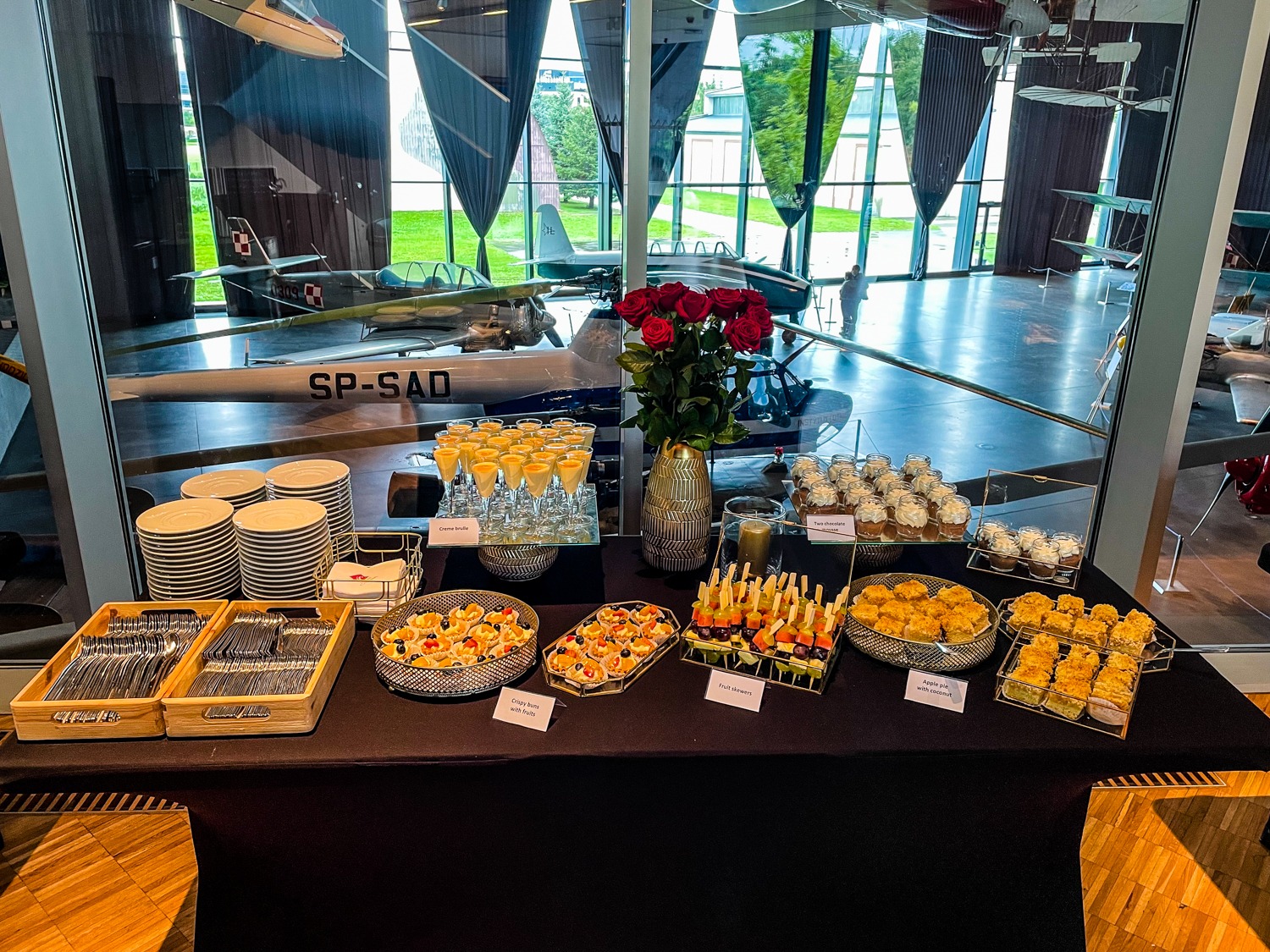 Stół cateringowy z deserami i przekąskami na czarnym obrusie, w tle ekspozycja samolotów w muzeum lotnictwa.