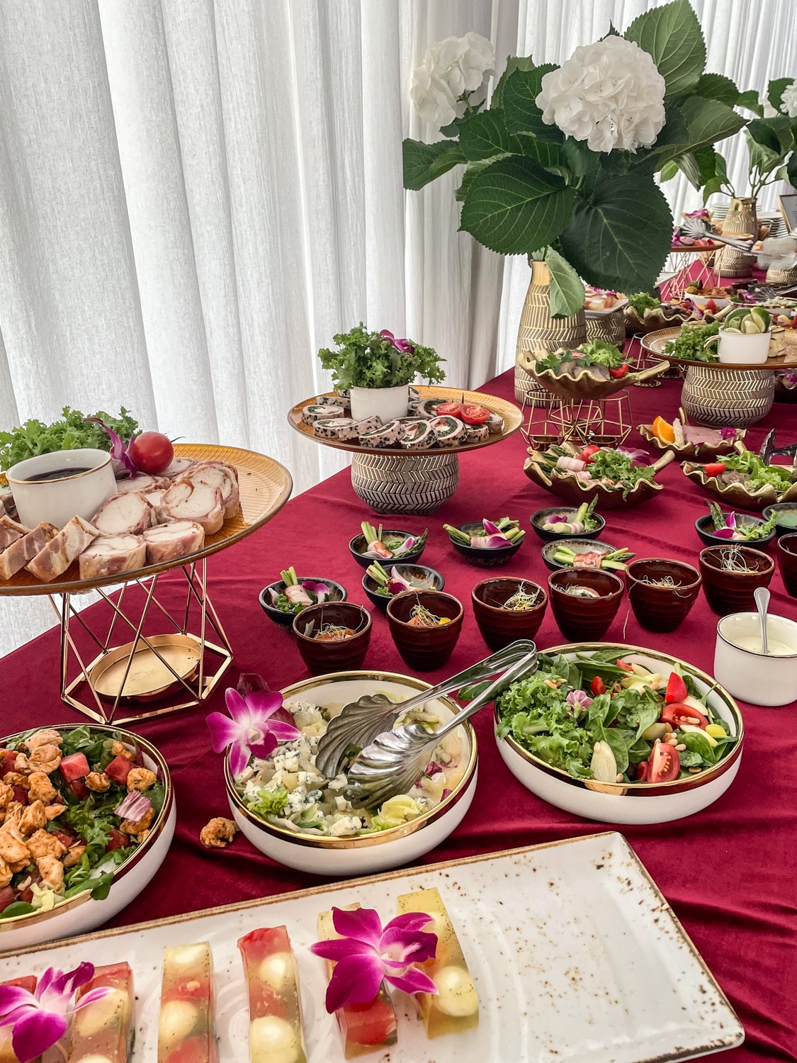 Bogato zastawiony stół z różnorodnymi sałatkami, roladkami mięsnymi, galaretkami i dekoracjami kwiatowymi na czerwonym obrusie, w tle białe zasłony i bukiet hortensji.