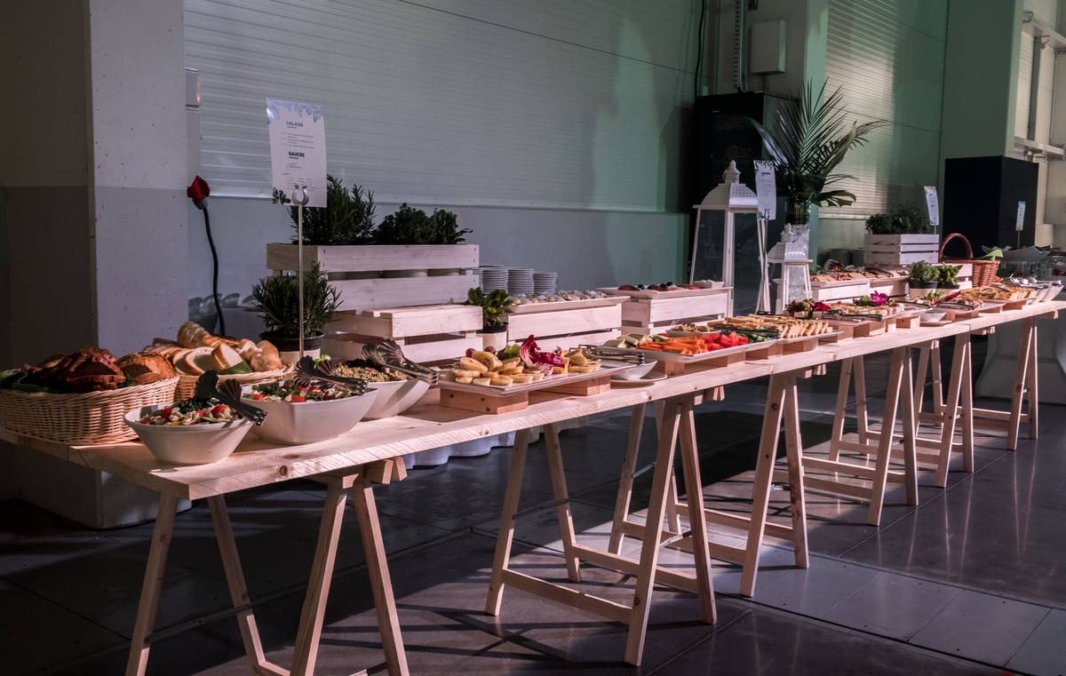 Długi stół cateringowy na drewnianych kozłach, zastawiony różnorodnymi przekąskami, sałatkami, pieczywem w koszykach, dekoracjami roślinnymi i białymi lampionami, z menu na stojaku w tle.
