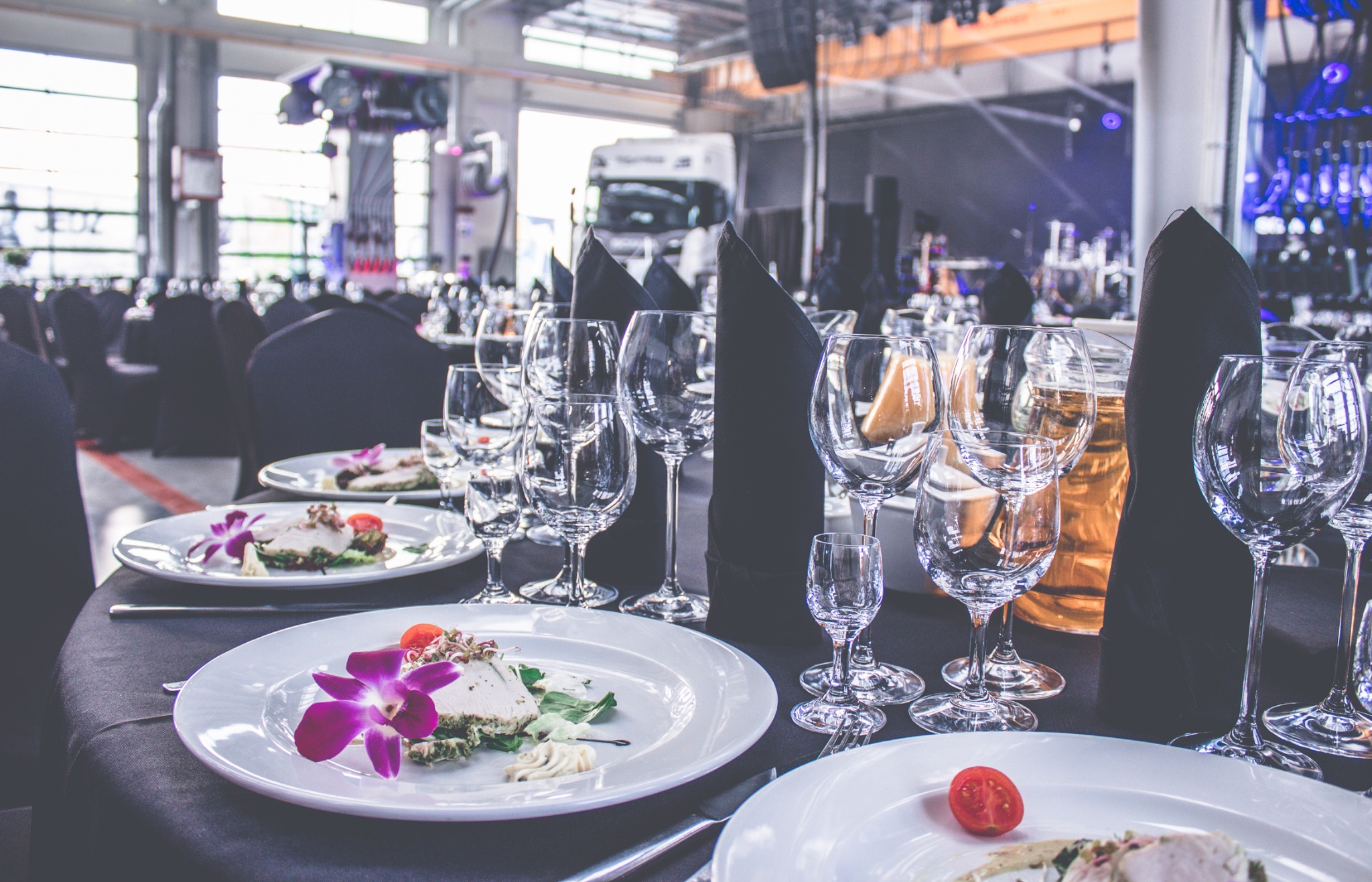 Elegancko nakryty stół z białymi talerzami, daniami z dekoracją kwiatową, licznymi kieliszkami i czarnymi serwetkami, w tle scena z oświetleniem i ciężarówka dostawcza.