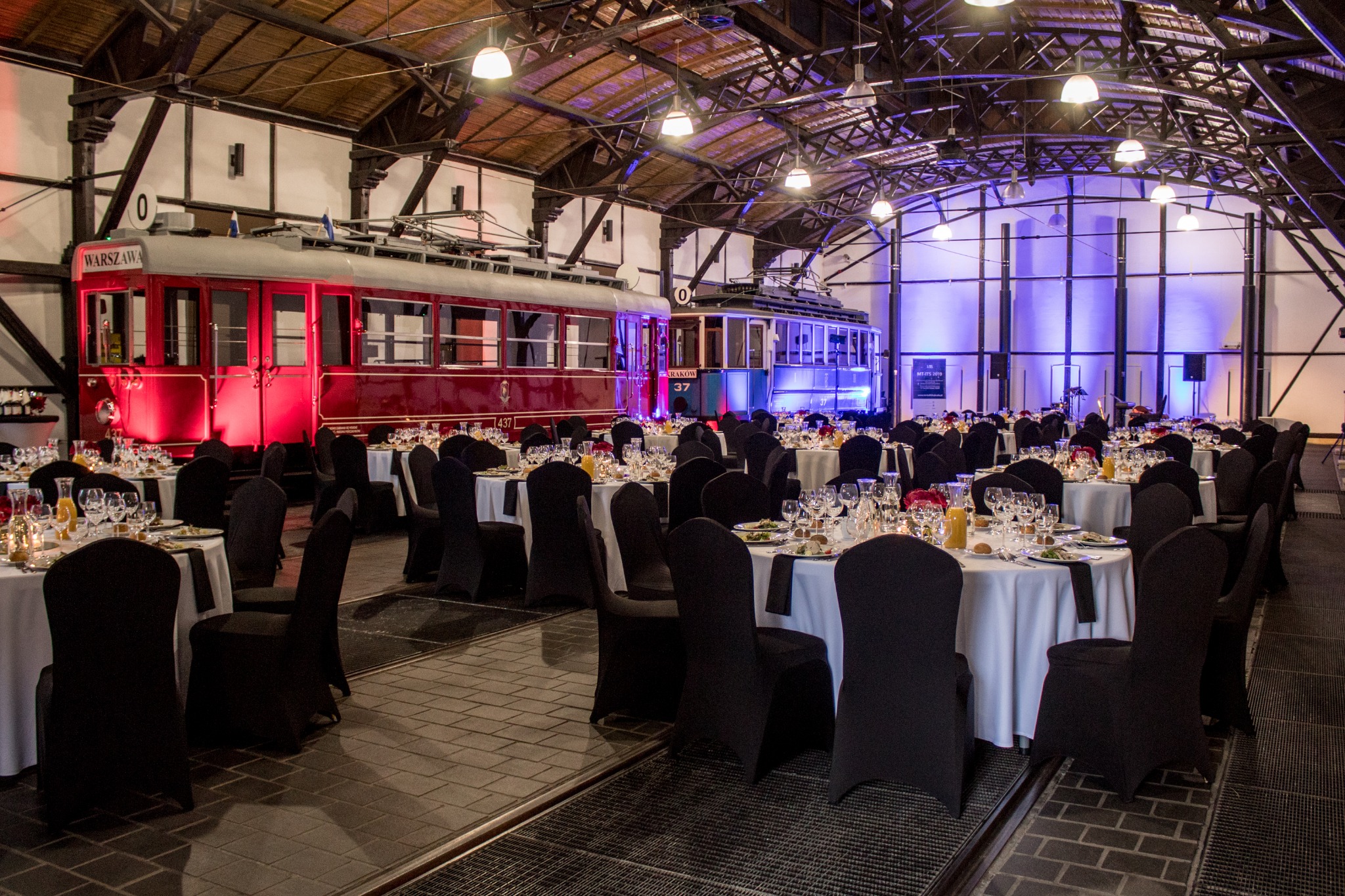Uroczysta sala bankietowa z okrągłymi stołami, nakryta dla gości, w tle zabytkowe tramwaje podświetlone na czerwono i niebiesko, industrialne wnętrze hali.