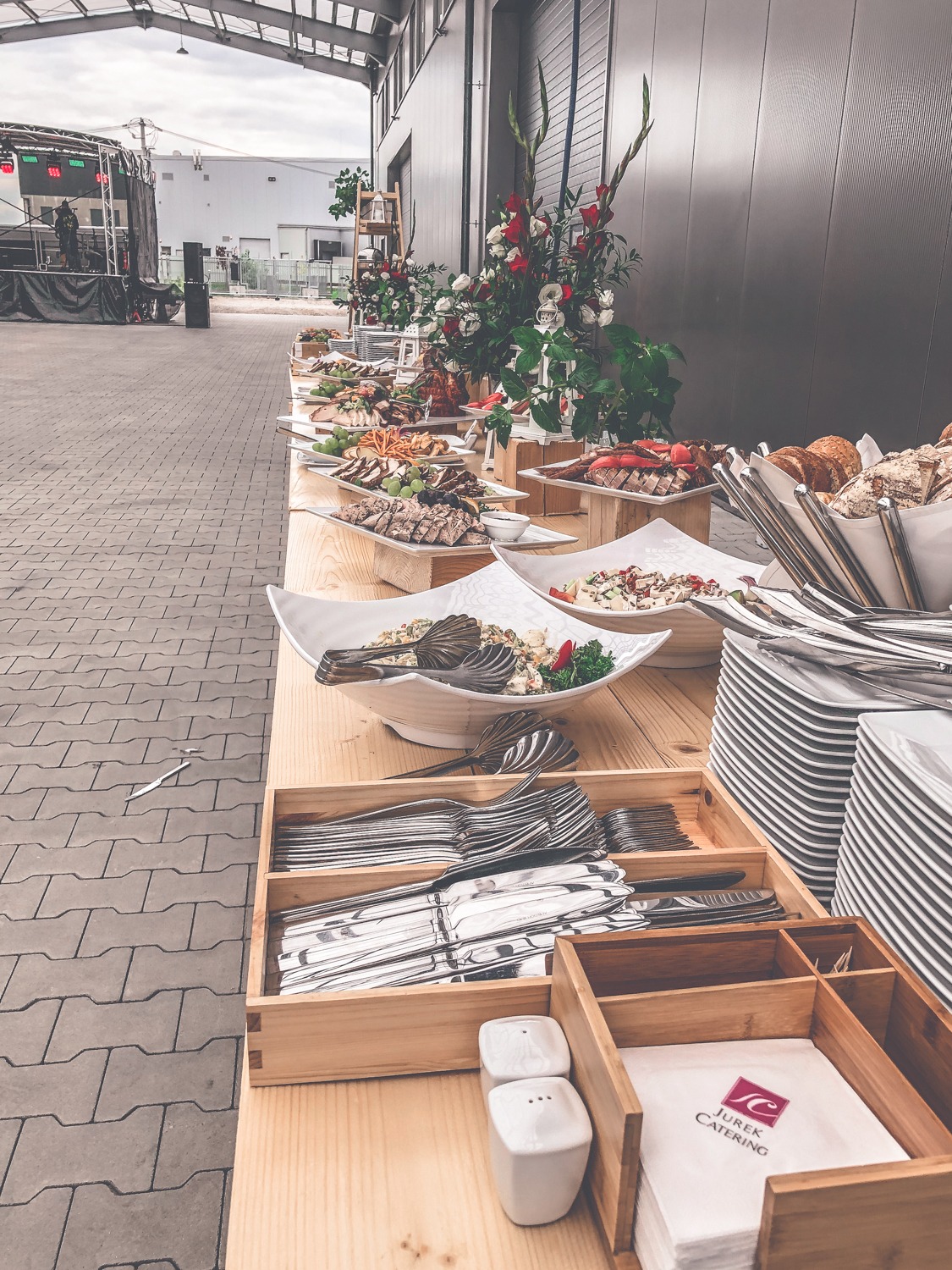 Stół cateringowy z przekąskami, mięsami, sałatkami, pieczywem, sztućcami i dekoracjami kwiatowymi na tle sceny koncertowej, papilotki z logo firmy cateringowej Jurek Catering.