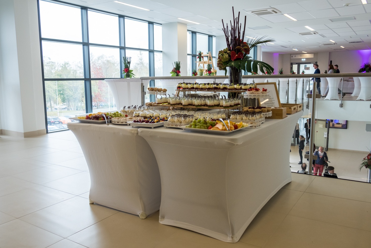 Stół z białym obrusem ugina się pod ciężarem różnorodnych przekąsek, owoców i deserów w szklanych naczyniach, wysoka kompozycja kwiatowa z ciemnych gałązek i czerwonych kwiatów w wazonie, tło...