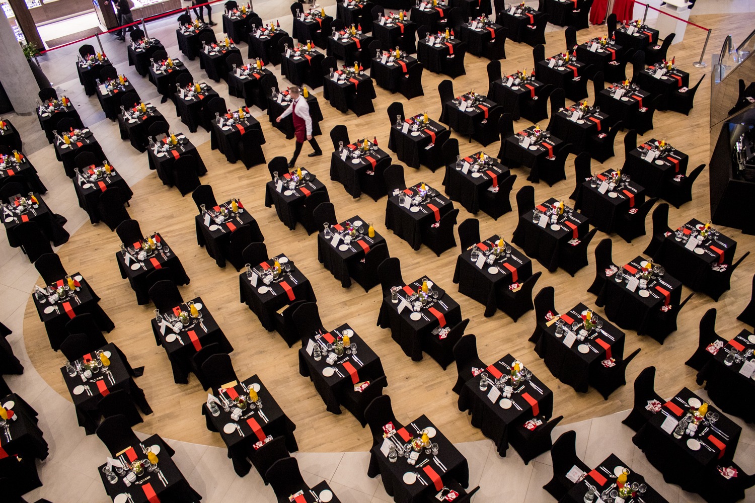 Widok z góry na salę balową z licznymi, identycznie nakrytymi stołami w czarnych obrusach z czerwonymi akcentami, przygotowanymi dla gości, kelner w oddali.