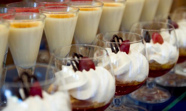 Rząd deserów w szklanych naczyniach: kremowe desery w wysokich kieliszkach obok deserów z bitą śmietaną, wiśnią i czekoladową dekoracją w szerszych kieliszkach.