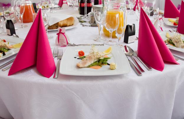 Elegancko nakryty stół z białym obrusem w delikatne wzory, różowe serwetki złożone w trójkąty, danie rybne na białym kwadratowym talerzu, szklanki i dzbanki z sokiem w tle.