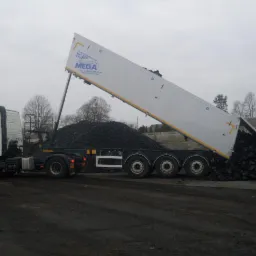 Ciężarówka marki MEGA z podniesioną naczepą wysypuje węgiel na plac składowy, widoczne drzewa w tle.
