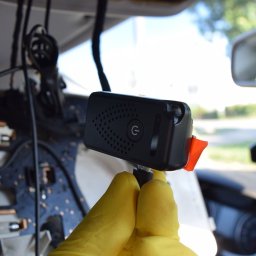 DETEKTYWI Agencja Detektywistyczna Gambit - Montaż kamery szpiegowskiej w samochodzie przez osobę w żółtych rękawiczkach, widoczne okablowanie i deska rozdzielcza. Detekcja techniczna.