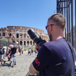 DETEKTYWI Agencja Detektywistyczna Gambit - Mężczyzna z długim obiektywem fotografuje Koloseum w Rzymie, widok zza krat, w tle spacerujący turyści i błękitne niebo.