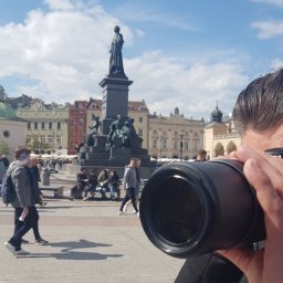 DETEKTYWI Agencja Detektywistyczna Gambit - Detektyw z aparatem na krakowskim rynku głównym, robiący zdjęcia. W tle pomnik Adama Mickiewicza i kolorowe kamienice. Słoneczny dzień.