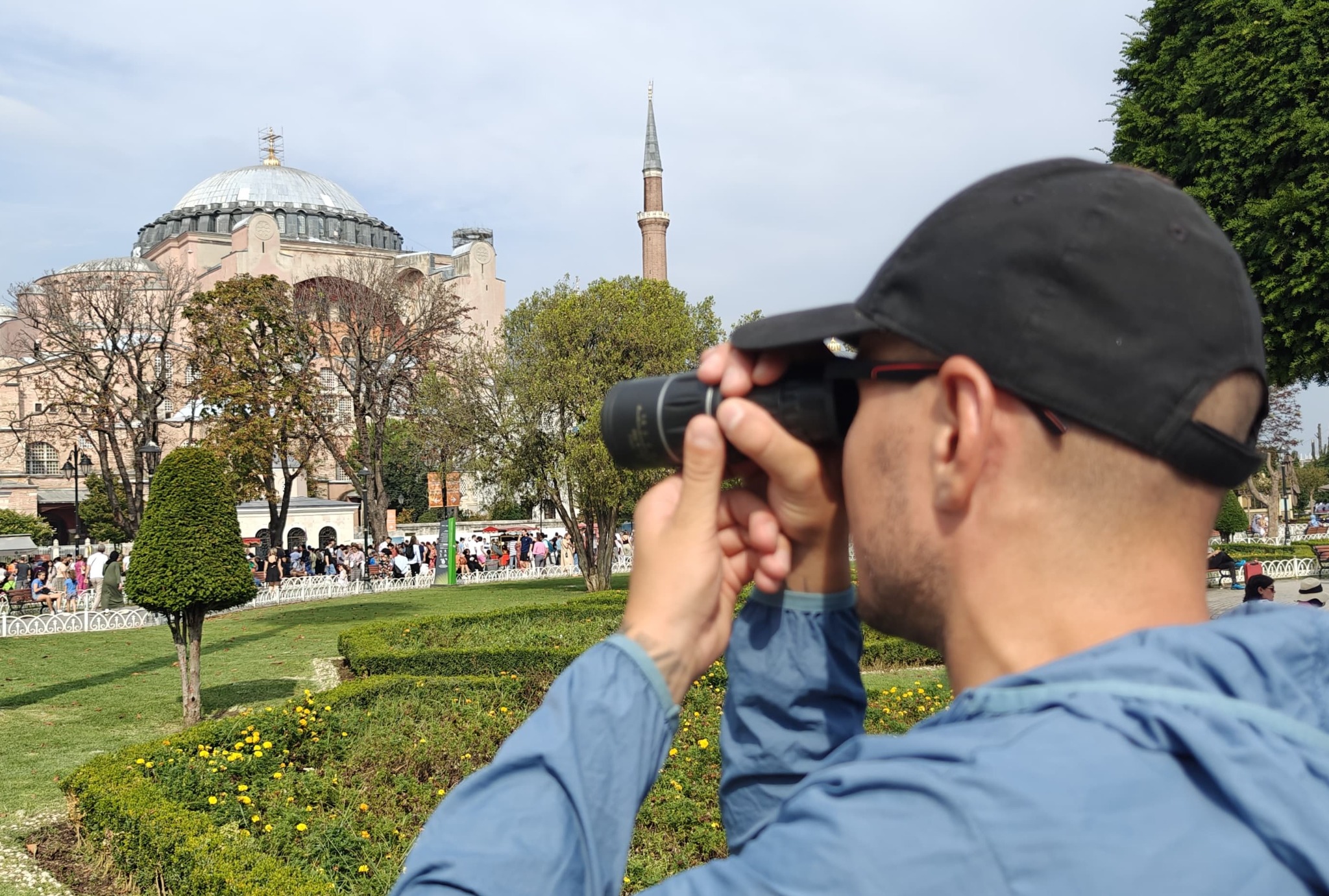 Mężczyzna w czapce obserwuje Hagia Sophia przez lunetę w słoneczny dzień. Turyści w tle, park z żółtymi kwiatami na pierwszym planie.
