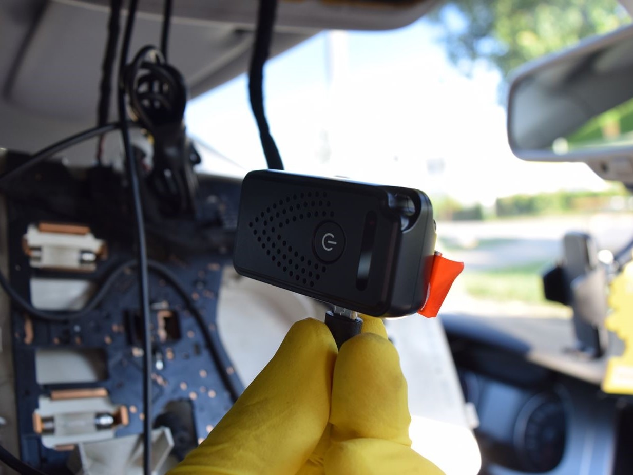 Montaż kamery szpiegowskiej w samochodzie przez osobę w żółtych rękawiczkach, widoczne okablowanie i deska rozdzielcza. Detekcja techniczna.