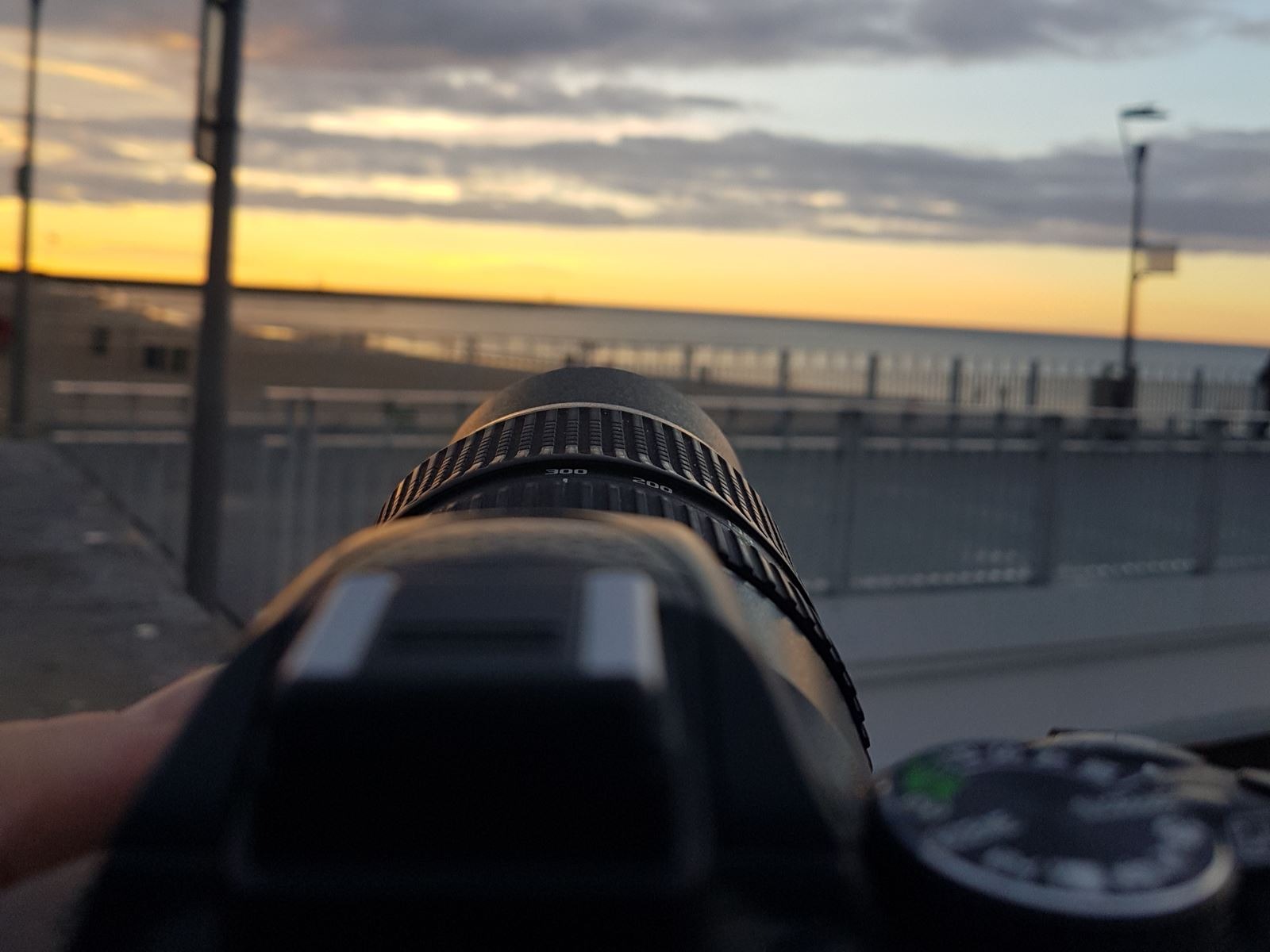 Obiektyw aparatu skierowany w stronę morza o zachodzie słońca, z widocznym fragmentem dłoni trzymającej sprzęt. Ujęcie z perspektywy fotografa.