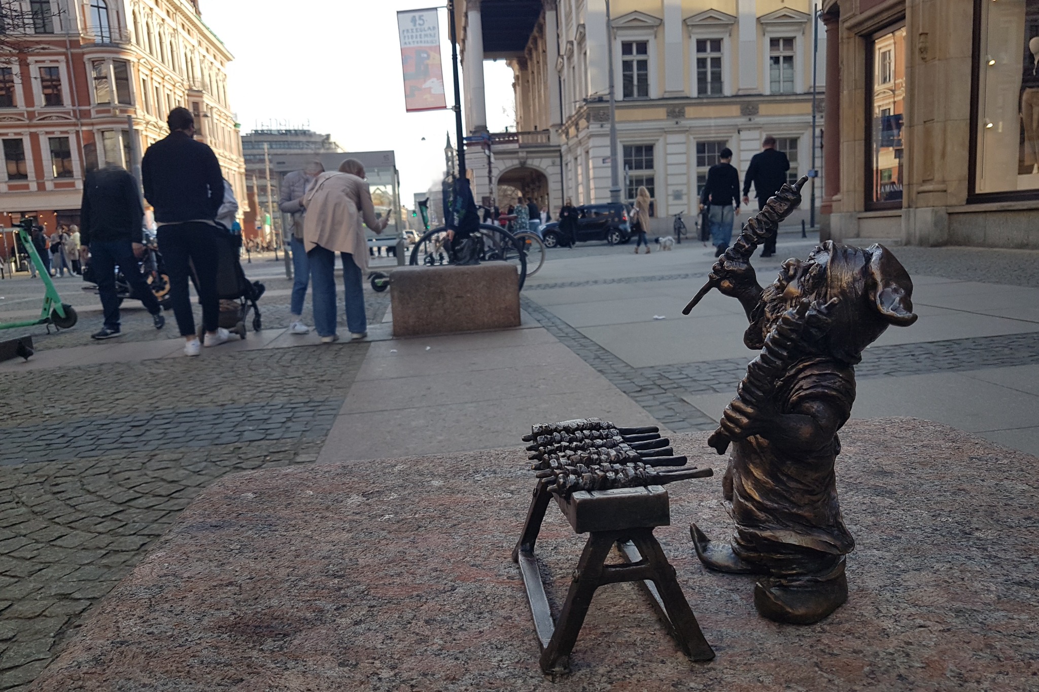 Brązowa figurka krasnala z rożnem i grillem na tle wrocławskiego rynku, w tle przechodzący ludzie i budynki. Nietypowe ujęcie z bliska.