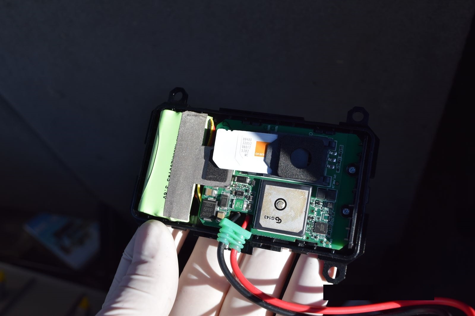 Otwarta obudowa lokalizatora GPS z kartą SIM Orange, baterią i płytką drukowaną, trzymana w dłoni w białej rękawiczce. Widoczne przewody zasilające.