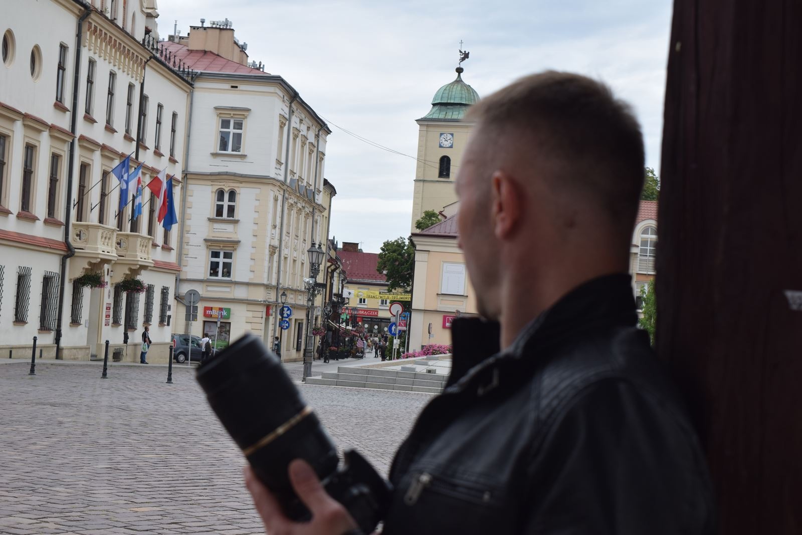 Osoba z aparatem w Lublinie, obserwująca rynek. Ujęcie zza ramienia, w tle budynki z flagami i wieża ratuszowa. Fotografia z perspektywy detektywa.