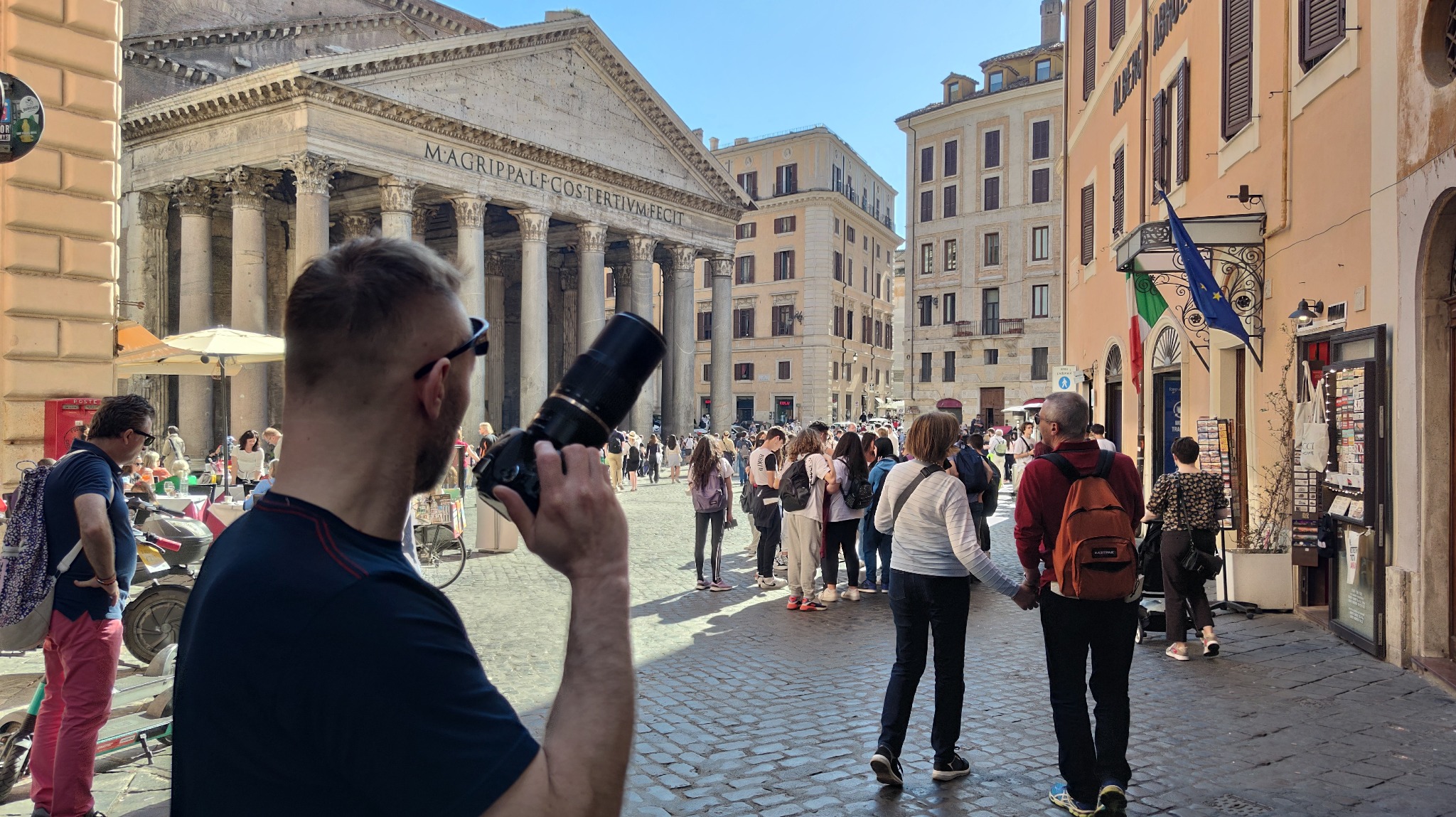Fotograf z aparatem na tle Panteonu w Rzymie, otoczony turystami. Ujęcie z bliska na fotografa i jego sprzęt, z Panteonem w tle. Słoneczny dzień.