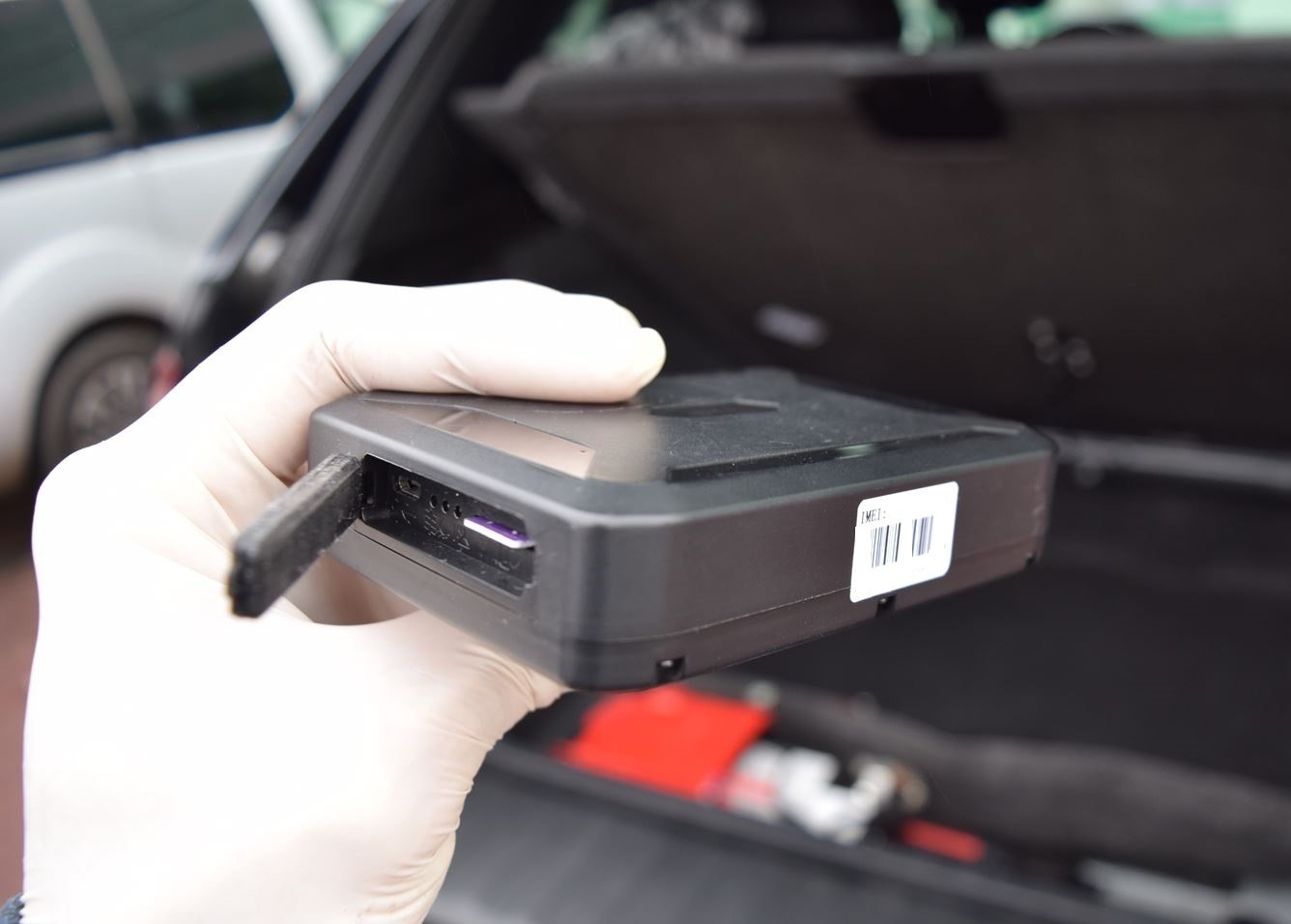 Czarna skrzynka z otwartą klapką na kartę SIM trzymana w rękawicy, widoczny kod kreskowy na etykiecie, tło bagażnika samochodowego, sugeruje monitoring.