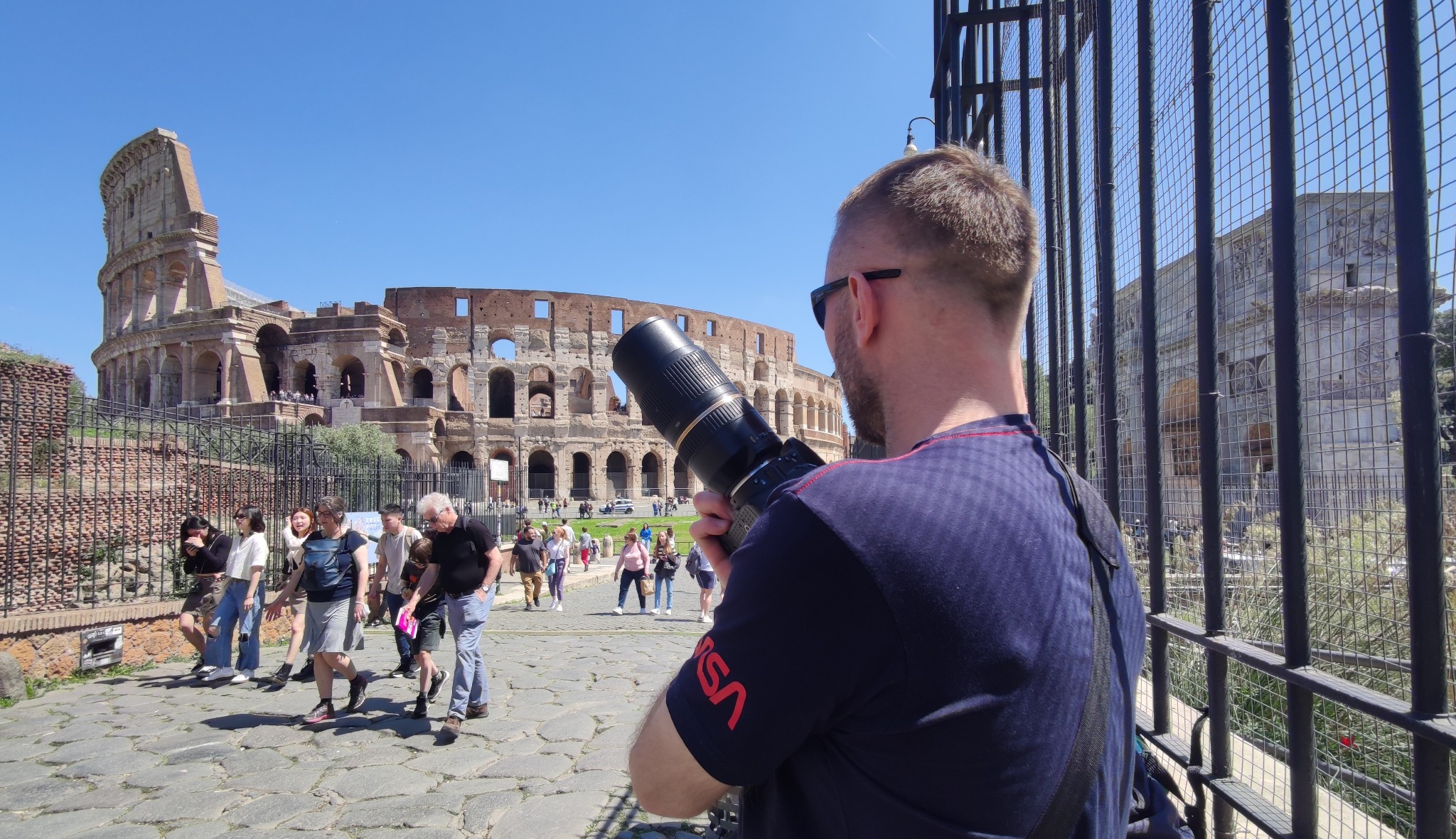 Mężczyzna z długim obiektywem fotografuje Koloseum w Rzymie, widok zza krat, w tle spacerujący turyści i błękitne niebo.