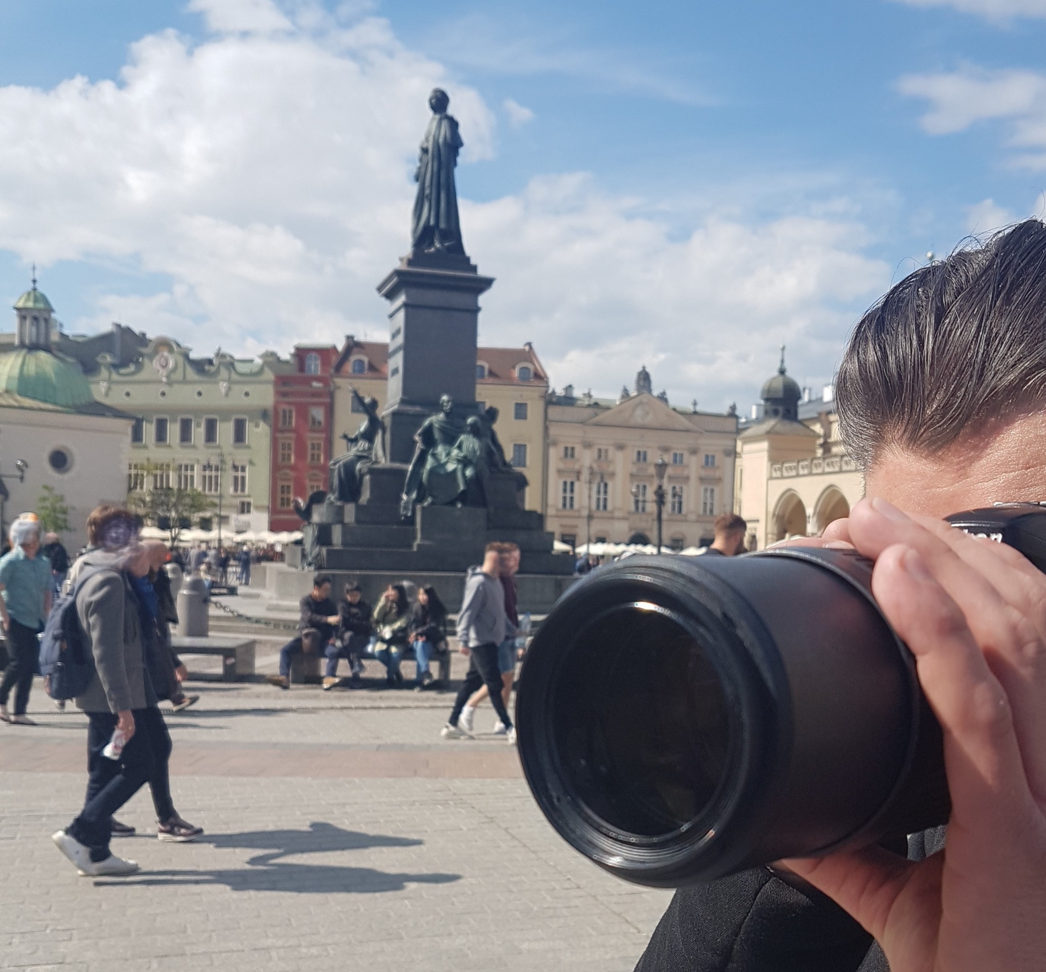 Detektyw z aparatem na krakowskim rynku głównym, robiący zdjęcia. W tle pomnik Adama Mickiewicza i kolorowe kamienice. Słoneczny dzień.