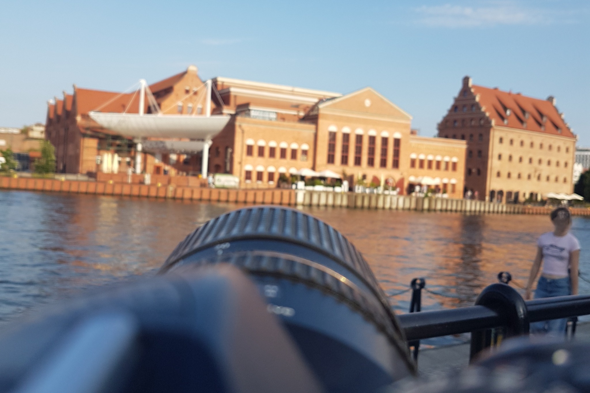 Widok na Motławę w Gdańsku z perspektywy obiektywu aparatu, w tle budynki Starego Miasta i spacerująca osoba. Rozmycie obiektywu na pierwszym planie.