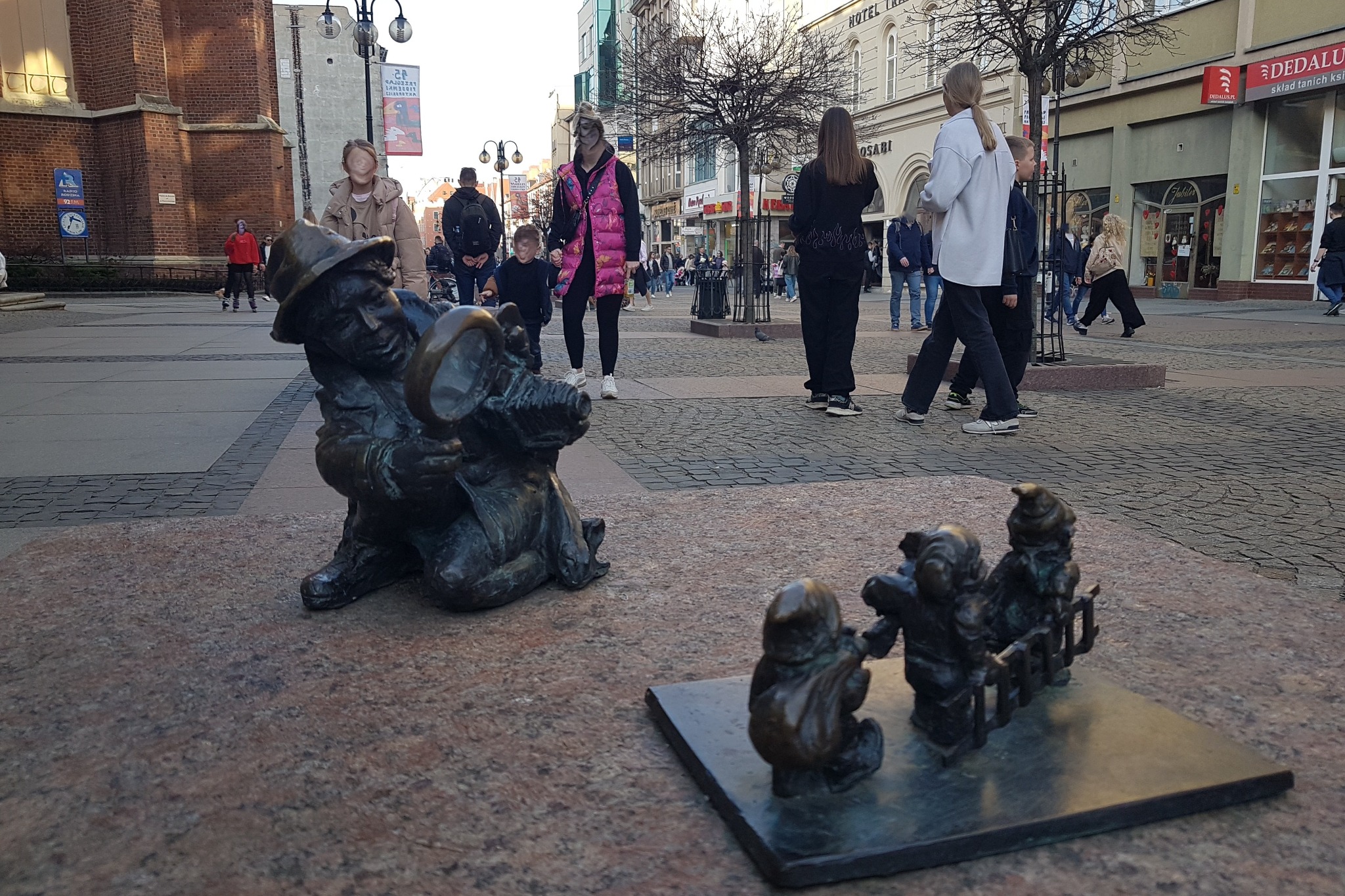 Brązowa figurka krasnala w kapeluszu, z lupą, na tle miejskiego placu z przechodniami i budynkami. W tle widoczny budynek Hotelu Osabi.