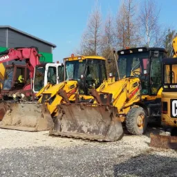 Rząd maszyn budowlanych: żółty Case, czerwony Atlas, dwie żółte koparko-ładowarki JCB 3CX i żółty mini-koparka Caterpillar CAT, stojące na żwirowym placu przed budynkiem z zielonym akcentem.