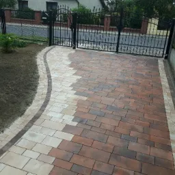 Nowo ułożony podjazd z kostki brukowej w odcieniach brązu i beżu, z dekoracyjnym obramowaniem, prowadzący do czarnej, metalowej bramy wjazdowej.