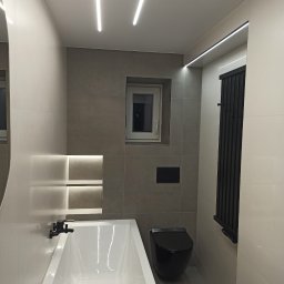 FHU Karol Formela - Nowoczesna łazienka z wanną, podwieszaną toaletą w kolorze czarnym i grzejnikiem drabinkowym. Oświetlenie LED w suficie i wnęce nad wanną. Szare płytki.