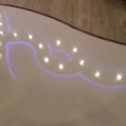 Jasne, błyszczące płytki podłogowe z dekoracyjnym podświetleniem LED w kształcie falistych linii, odbijające światło punktowych lamp, z widoczną krawędzią drewnianej podłogi w tle.