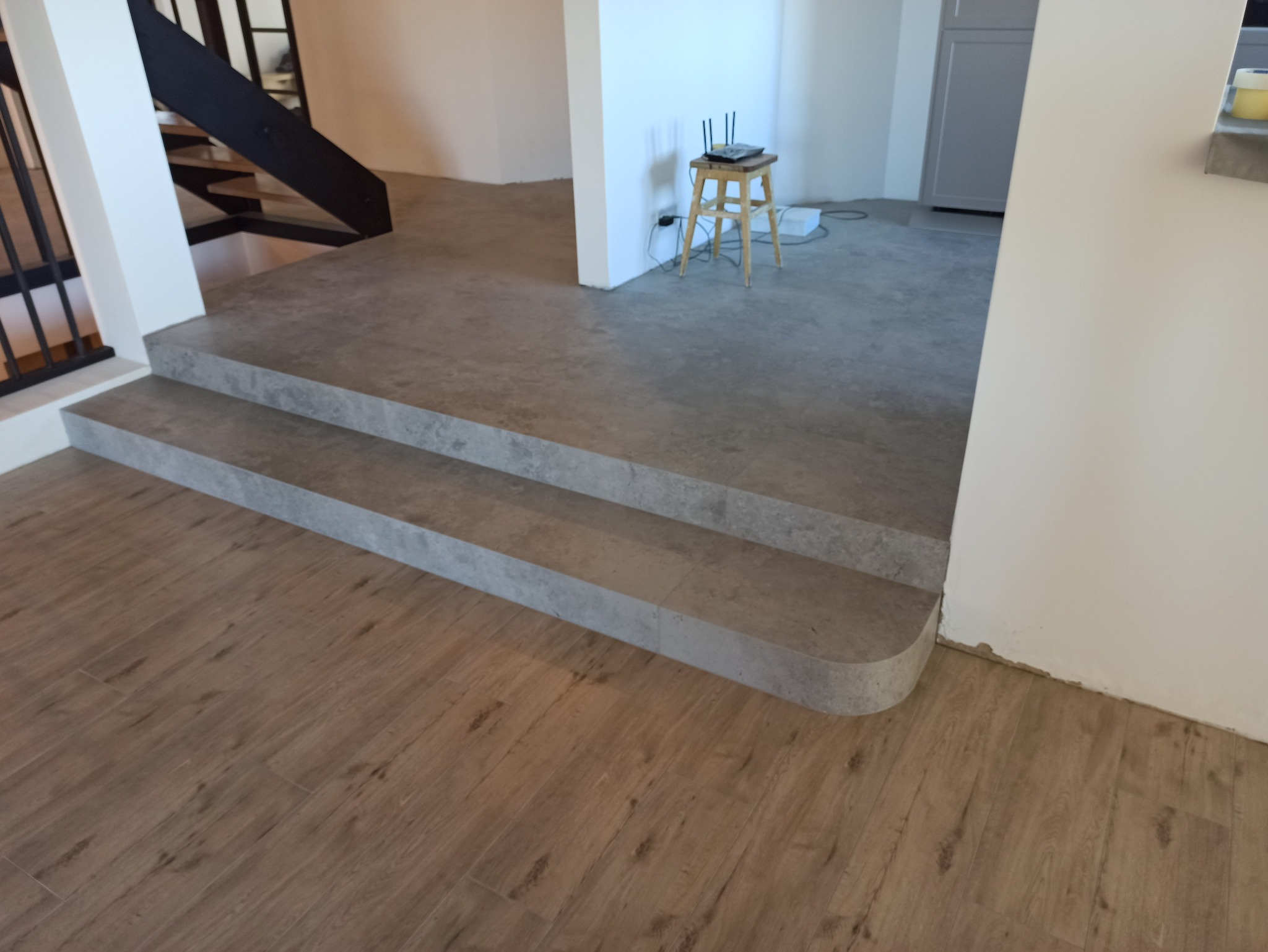 Wykończone schody z szarej płytki imitującej kamień, prowadzące do pomieszczenia z drewnopodobną podłogą. Widoczny fragment metalowej konstrukcji schodów.