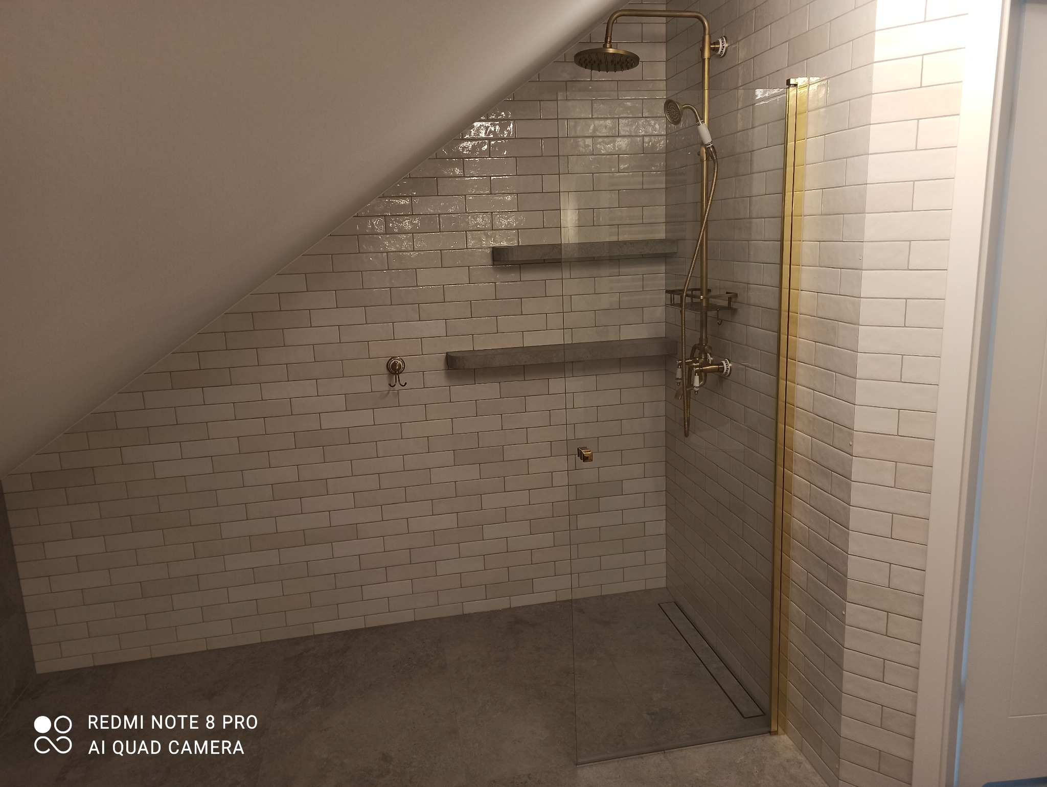 Ukończona łazienka na poddaszu ze szklaną kabiną prysznicową, wykończona białymi, błyszczącymi płytkami imitującymi cegłę i złotą armaturą. Widoczne półki.