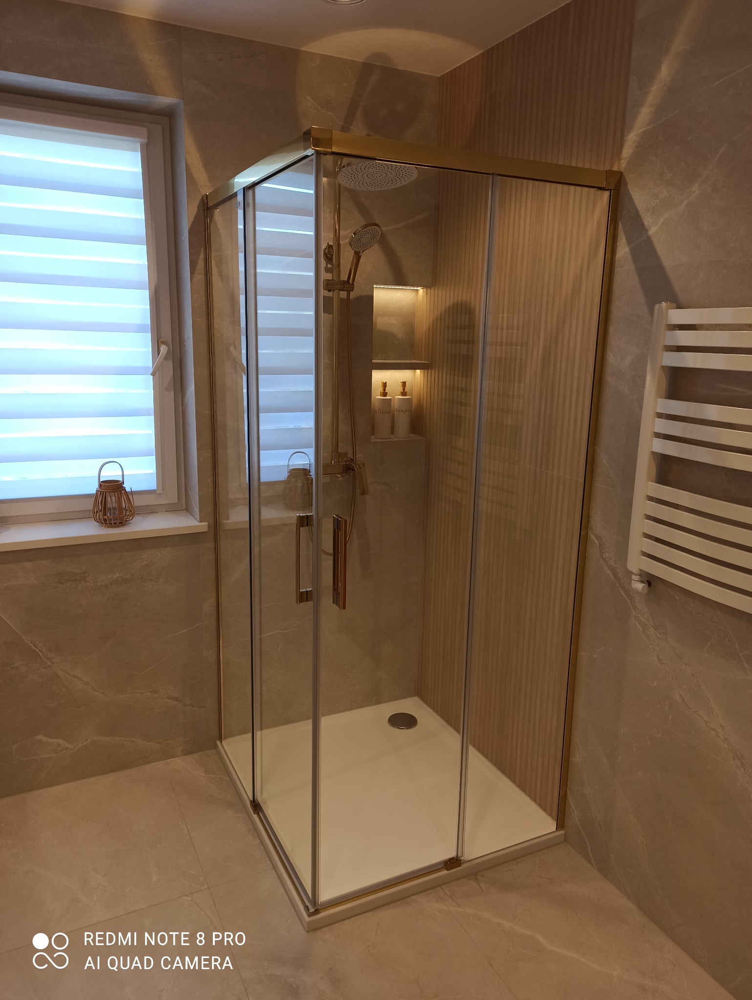 Nowoczesna kabina prysznicowa z złotymi detalami, jasne płytki imitujące marmur, grzejnik drabinkowy i okno z żaluzjami w tle. Eleganckie wykończenie łazienki.